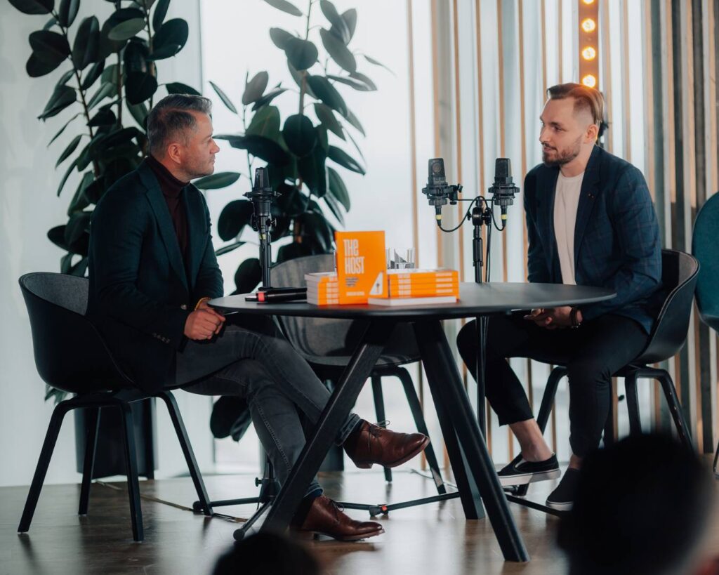 Jarosław Kuźniar podczas wywiadu w studiu podcastowym z książką The Host na stole, dwóch mężczyzn siedzących przy profesjonalnych mikrofonach w nowoczesnym wnętrzu z roślinami