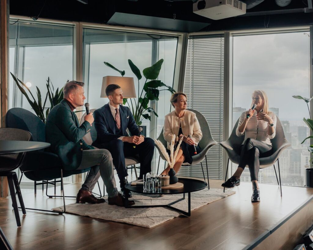 Panel dyskusyjny z Jarosławem Kuźniarem i trzema uczestnikami w nowoczesnym biurowcu z widokiem na miasto, prowadzący z mikrofonem podczas premiery podręcznika The Host