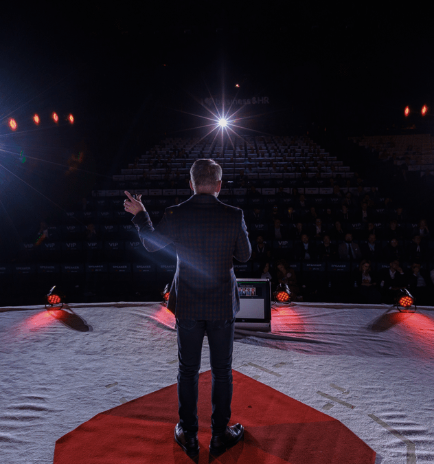 Jarosław Kuźniar na scenie prowadzi przemówienie – widok zza pleców, sala pełna słuchaczy